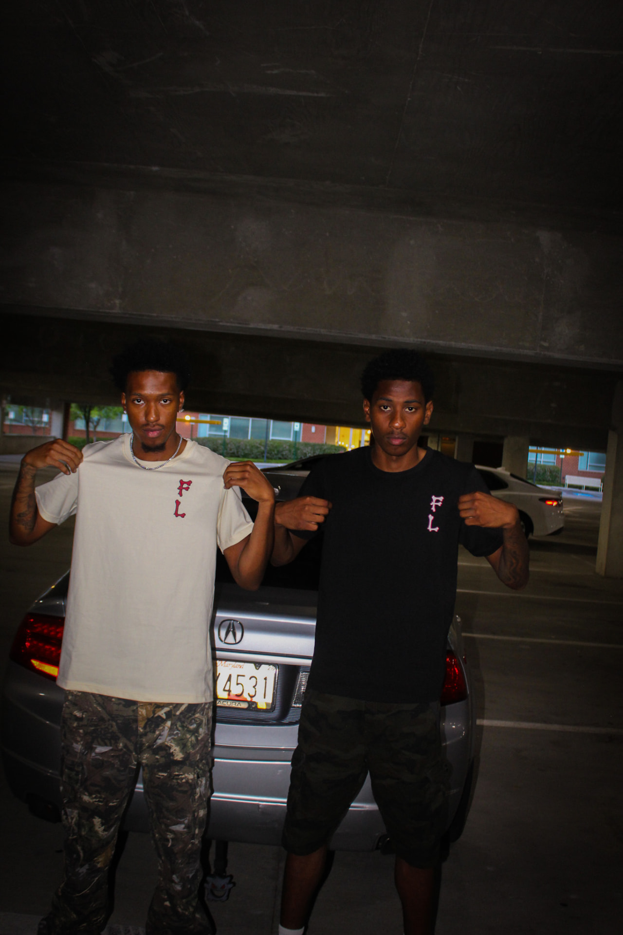 Two models showing off black and crème F8VRLASTING tees in a parking garage